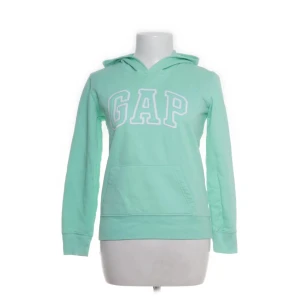 Mintgrön hoodie från GAP - Fräsch mintgrön hoodie från GAP med stor vit logga framtill. Tröjan har huva, magficka och långa ärmar. Perfekt för chill dagar och enkel att matcha med jeans eller leggings. Mjuk bomullskvalitet och klassisk passform.