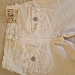 Vita bootcut jeans från True Religion - Säljer ett par vita True Religion jeans som är bootcut som är lågmidjade. Innerbenslängden är 80cm och midjemåttet rakt över 36cm. Nyköpta kostar 1300 kr
