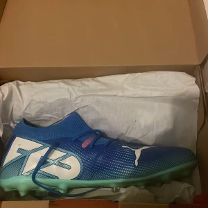 Puma Future 7 blå fotbollsskor strl 43 - Säljer ett par Puma Future 7 Match MG fotbollsskor i blått med vita och mintgröna detaljer. Skorna har stickad strumpkrage, snörning och en platt sula med dobbar för gräsplan. Snygg design med stor Puma-logga på sidan och coola grafiska inslag. Original pris 1100