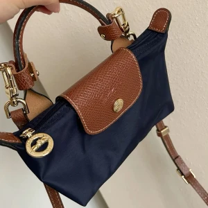 Blå och brun handväska från Longchamp - Snygg handväska från Longchamp i marinblått tyg med bruna detaljer i skinn. Väskan har guldfärgade metalldetaljer, ett klassiskt lock med tryckknapp och både handtag och axelrem i brunt skinn. Perfekt storlek för det viktigaste och stilren design.