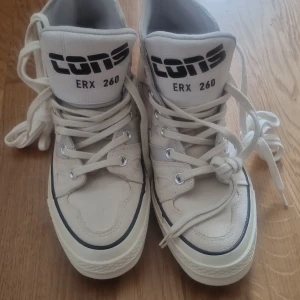 Converse Cons ERX 260 vita high-top sneakers - Säljer ett par Converse Cons ERX 260 high-top sneakers i vit färg med svarta detaljer och logga. Skorna har snörning, rund tå och platt sula. Materialet är en mix av canvas och syntet, med extra vaddering runt ankeln för skön komfort. Perfekt för dig som gillar retrostil och streetwear.