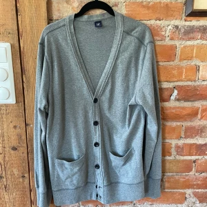 Grå kofta från GAP med knappar - Snygg grå kofta från GAP i storlek L. Koftan har v-ringad hals, svarta knappar framtill och två praktiska fickor. Tillverkad i mjuk bomull, perfekt för lager-på-lager. Långärmad och stilren design som funkar till många outfits.