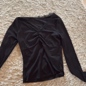 Svart långärmad topp med spets - Svart långärmad topp med snygg v-ringning och spetsdetaljer runt halsen. Toppen har en lätt rynkning framtill som ger en cool form och är gjord i mjukt, stretchigt material. Perfekt för dig som gillar stilrena och trendiga plagg. Oanvänd i strl S.
