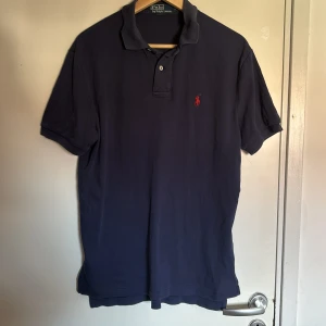Marinblå Polo t-shirt från Ralph Lauren - Snygg marinblå Polo t-shirt från Ralph Lauren med klassisk krage och två knappar framtill. Ikonisk röd broderad logga på bröstet. Tillverkad i mjuk bomull som är skön mot huden. Perfekt för dig som gillar stilren och tidlös design.