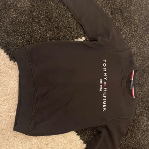 Svart sweatshirt från Tommy Hilfiger - Svart sweatshirt från Tommy Hilfiger med rund hals och tryckt logga på bröstet. Tröjan har ribbade muddar vid ärmslut och nederkant samt klassisk passform. Materialet är mjuk bomull och polyester, perfekt för chill dagar.