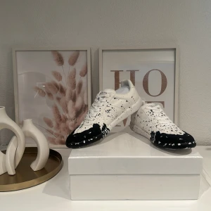 Maison Margiela sneakers svart/vit - Unika sneakers från Maison Margiela i vitt skinn med svart färgstänksmönster över tå och sidor. Klassisk gummisula i brunt och vit ovandel med snörning. Snygg och edgy design som sticker ut, perfekt för dig som gillar streetstyle och konstnärliga detaljer.