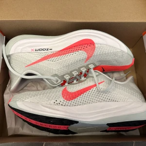 Nike zoomx Pegasus turbo plus vit/rosa - Nike ZoomX  pegasus turbo plus sneakers i ljusgrått med starkt rosa detaljer och swoosh. Ovandelen är ventilerande mesh, snörning och platt sula med svart och rosa grepp. Perfekt för dig som vill maxa din löpning med stil och komfort.