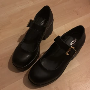 Svarta Mary Jane-plattformskor från Koi - Säljer ett par svarta Mary Jane-plattformskor från Koi med chunky klack och grov sula. Skorna har rund tå, bred rem med guldfärgat spänne och är tillverkade i syntetmaterial. Perfekt för dig som gillar statement-skor med retrovibe och extra höjd.