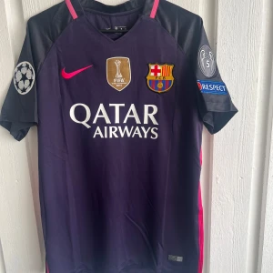 FC Barcelona Neymar Jr 11 matchtröja - Säljer en FC Barcelona matchtröja med Neymar Jr och nummer 11 på ryggen. Tröjan är mörklila med svarta detaljer och rosa tryck, har korta ärmar och Champions League-patch på båda ärmarna. Materialet är lätt och andas, perfekt för fotboll eller samling.