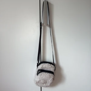 Vit fluffig axelväska med dragkedja - Supermysig vit axelväska i teddy-material med två fack och svarta dragkedjor. Perfekt storlek för mobilen och småsaker, och har en justerbar svart axelrem. Väskan har en rundad form och ger en cool vibe till din outfit.