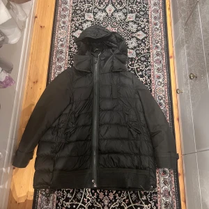 Grön dunjacka från Moncler - Säljer en svart dunjacka från Moncler med huva och quiltad design. Jackan har dragkedja framtill, knappar och praktiska fickor. Materialet är polyester med dunfyllning, perfekt för kalla dagar. Snygg och stilren look som funkar till många outfits. Mycket bra skick endast små hål och en saknad knapp