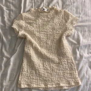 Krämvit blus från Kappahl XS - Supersöt krämvit blus från Kappahl i storlek XS. Den har korta ärmar, ett ribbat och strukturerat tyg med volangkant vid halsen. Perfekt för dig som gillar Sthlm stil och chill stil🪩. Materialet känns mjukt och luftigt, och färgen är ljus och fräsch.