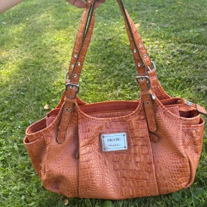 Supersnygg vintage väska❤️ - Snygg orange/ljusbrun handväska med krokodilpräglad yta från Nicole by Nicole Miller. Väskan har justerbara axelremmar med silverfärgade spännen och flera praktiska fack, inklusive dragkedja på baksidan. Perfekt för dig som gillar att sticka ut med coola detaljer. Sååååå såååå snygg. Köpt sacond hand. Fått så många komplimanger💕
