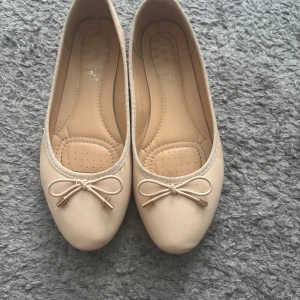 Beige ballerinaskor med rosett - Snygga beige ballerinaskor med rund tå och dekorativ rosett framtill med guldfärgade detaljer. Skorna har en mjuk och skön innersula och är tillverkade i syntetmaterial. 
