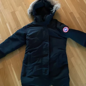 Svart dunparkas från Canada Goose - Säljer en svart dunparkas från Canada Goose med huva och snygg pälsdetalj. Jackan har lång ärm, klassisk logga på armen och praktiska fickor framtill. Perfekt för kalla dagar med sin varma dunfyllning och stilrena look. Säljs pågrund av att den inte används köptes för 6000 för 2 år sen