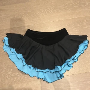 Svart och blå volangkjol från Jerry's - Snygg kortkjol från Jerry's med svart midja och dubbla volanger i svart och ljusblått. Kjolen har en riktigt flowig form som ger cool rörelse och är tillverkad i mjuk polyester med lite stretch. Perfekt för dig som gillar att sticka ut med färg och volanger.