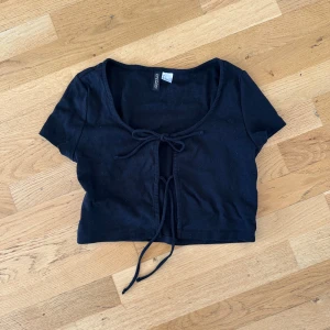 Svart croppad bolerotopp från Divided - Svart bolerotopp från Divided med korta ärmar och croppad passform. Toppen har snygg snörning framtill som knyts i två rosetter och en djup rundad urringning. Perfekt att styla över linnen eller klänningar för en trendig look.