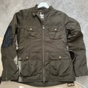 Olivgrön parkajacka med bälte - Snygg olivgrön parkajacka från Boomerang med fyra framfickor, dragkedja och knappar. Jackan har svart patch på ena ärmen och ett justerbart bälte i midjan. Perfekt för dig som gillar funktionell stil med coola detaljer.