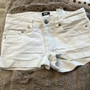 Vita jeansshorts från H&M Sure of Heart - Snygga vita jeansshorts från H&M med Sure of Heart-detalj på bakfickan. Klassisk femficksmodell med knapp och dragkedja framtill. Shortsen har en lågmidjad passform och är gjorda i bomull, perfekta för varma sommardagar.