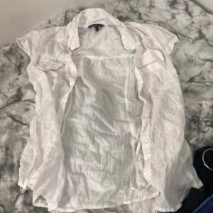 Vit randig skjorta från OVS Young - Vit skjorta med tunna ränder från OVS Young, storlek 40. Skjortan har korta ärmar, klassisk krage och knappar framtill. Materialet är lätt och luftigt, perfekt för varmare dagar. På ryggen finns en broderad fjäril i vitt som ger en extra detalj. Passar mig som Xs-S