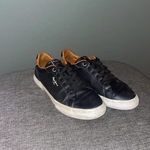 Svarta sneakers från Pepe Jeans - Snygga svarta sneakers från Pepe Jeans med bruna detaljer runt hälen och insidan. Skorna har snörning, rund tå och vit platt sula med Pepe Jeans-logga under. Materialet är skinn och syntet, perfekt för dig som gillar stilrena och klassiska sneakers.