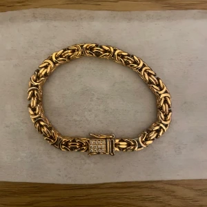 Guldigt armband med glittrig lås - Säljer ett chunky armband i guldton med flätad design och en låsdel dekorerad med små glittrande stenar. Armbandet har en cool och unik struktur som verkligen sticker ut och passar perfekt om du vill ha något som syns.