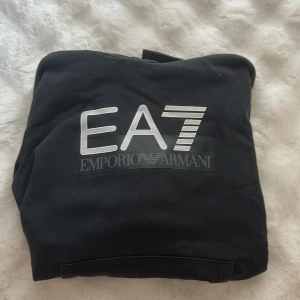 Svart EA7 Emporio Armani hoodie - Svart hoodie från EA7 Emporio Armani med stor logga i silver på bröstet. Tröjan har huva med snörning, ribbade muddar och magficka. Materialet är mjuk bomull och polyester, perfekt för chill dagar. Snygg och enkel design som funkar till jeans eller joggers.