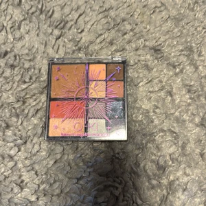 QKI sminkpalett med flera färger - Snygg sminkpalett från QKI med 10 olika nyanser för ansikte och ögon. Innehåller bronzer, rouge och ögonskuggor i varma toner som orange, brunt, guld, beige, silver och lila. Paletten har ett coolt stjärnmönster på locket och är kompakt och lätt att ta med.