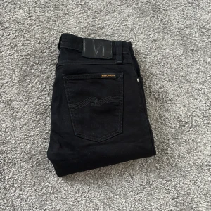 Nudie Jeans 28/32 - Svarta jeans från Nudie Jeans med klassisk femficksdesign och snygga svarta sömmar på bakfickan. Modellen har normal passform och raka ben, med silverfärgade nitar och knapp. Tillverkade i mjukt jeansmaterial som sitter skönt hela dagen.