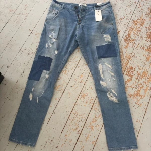 Blå patchade jeans från Zhrill - Snygga blå jeans från Zhrill med slitna detaljer och mörkare denimpatchar på benen. Modellen har raka ben, klassisk femficksdesign och knappar i gylfen. Jeansen är tillverkade i bomull och har en cool, streetig vibe med tydliga slitningar och lagningar.