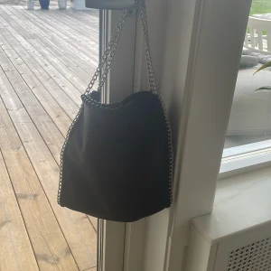 Svart axelväska med kedjedetaljer - Svart axelväska från Gina Tricot med silverkedja som axelrem och runt kanterna.  Insidan har en ficka med dragkedja och gott om plats för dina grejer.