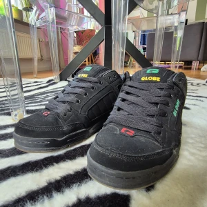 Globe Tilt (42)  - Sköna och robusta Globe Tilt sneakers i svart med färgdetaljer i gult, grönt och rött. Skorna har tjock platt sula, rund tå och klassisk snörning. Ovandelen är i skinn och syntet, med vadderad plös och häl för extra komfort. Perfekta för skate eller streetstyle.