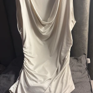 Vit omlott linnetopp från H&M - Säljer en vit linnetopp från H&M med snygg draperad halsringning och omlott-effekt. Toppen är ärmlös och har cool rynkning på sidan som ger en extra touch. Materialet är mjukt och har lite glans, perfekt för att lyfta din outfit.