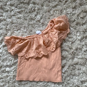 Persikofärgad offshoulder topp med volang - Superfin persikofärgad offshoulder topp från Zara  med bred volangkant och broderade blommor. Toppen är ärmlös och har en elastisk kant som gör att den sitter snyggt på axlarna. Perfekt för sommaren🩷