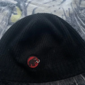 Svart mössa från Mammut - Snygg svart mössa från Mammut med deras ikoniska logga i rött framtill. Mössan är ribbad och perfekt för kyliga dagar. Den har en diskret etikett på sidan och är fodrad för extra värme.