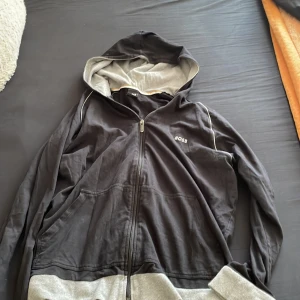 Svart huvtröja från Hugo Boss - Säljer en svart huvtröja från Hugo Boss med dragkedja och grå detaljer. Tröjan har långa ärmar och en bekväm passform. Perfekt för en avslappnad stil. 🖤