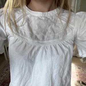 Vit blus med broderi - Säljer en söt vit blus med korta ärmar och vackert broderi på bröstet. Blusen är croppad och har en luftig passform, perfekt för varmare dagar. Den har en rund halsringning och fina detaljer som ger en romantisk känsla. Den har ett sprucket hål i ryggen som inte syns så mkt. 