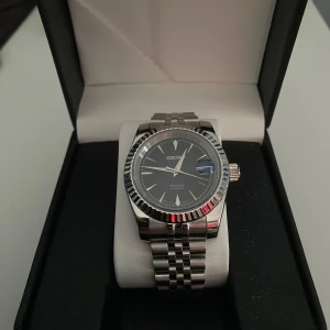 Seiko klocka - Elegant Seiko-klocka med silverfärgad boett och armband i rostfritt stål. Den har en mörkblå urtavla med datumvisning och tydliga markeringar. Klockan har en automatisk rörelse och en transparent baksida som visar mekanismen. Perfekt för den som uppskattar stil och funktion.