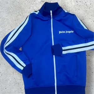 Blå track jacket från Palm Angels - Snygg blå track jacket från Palm Angels med vita och svarta ränder längs ärmarna. Jackan har en dragkedja framtill och en hög krage. Perfekt för en sportig look!