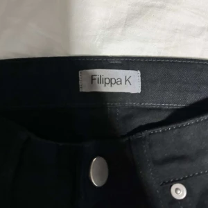 Svarta jeans från Filippa K - Snygga svarta jeans från Filippa K med klassisk design och knappstängning. Byxorna har fem fickor och är tillverkade i ett bekvämt material som passar perfekt för en stilren look.
