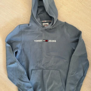 Blå hoodie från Tommy Jeans - Säljer en snygg blå hoodie från Tommy Jeans med klassisk logga på bröstet. Tröjan har en bekväm passform med långa ärmar och en praktisk magficka. Som ny, strl xs. Rök och djurfritt hem.