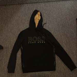 Svart hoodie från Hugo Boss - Snygg svart hoodie från Hugo Boss med guldfärgad logga på framsidan. Hoodien har en justerbar huva med beige foder och långa ärmar. Perfekt för en stilren och bekväm look.