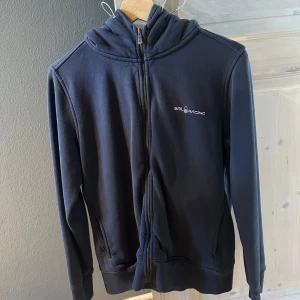 Mörkblå hoodie från Sail Racing - Säljer en mörkblå hoodie från Sail Racing med dragkedja framtill. Tröjan har en broderad logga på bröstet och en liten logga på huvan. Perfekt för en avslappnad stil.