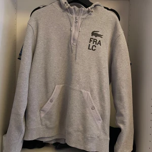Grå Lacoste Hoodie - Säljer en grå halfzip hoodie från Lacoste FRA N43 kollektion. Tröjan har en  logga på bröstet och text på ärmen. Även en logga och text på sidan av luvan.