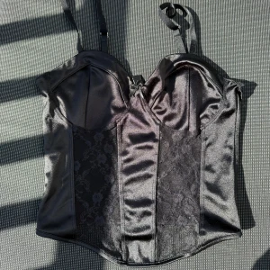 Svart korsett med spetsdetaljer - Snygg svart korsett från H&M med spetsdetaljer och justerbara axelband. Perfekt för en stilfull look. Korsetten har en glansig finish och en elegant design.