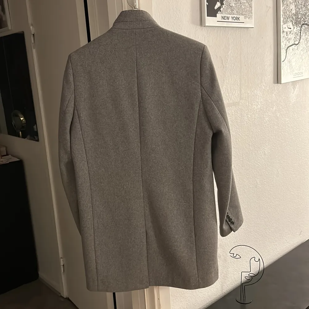 Snygg grå kappa med stilrena knappar framtill. Tillverkad i en ullblandning för en elegant look. Perfekt för kyligare dagar med sin långa design och praktiska fickor.. Takit.
