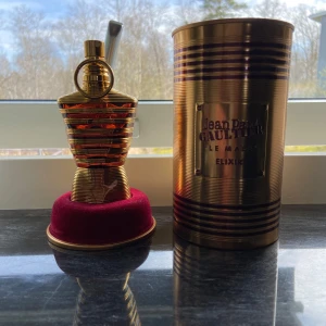 Jean Paul Gaultier Le Male Elixir - Exklusiv parfymflaska i form av en manlig torso med guld- och bronsfärger. Flaskan står på en röd sammetssockel och kommer med en matchande metallcylinder som förpackning. Perfekt för den som vill ha en lyxig och unik doftupplevelse. 55-60ml kvar av 75