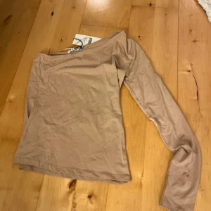 Beige one shoulder topp från NA-KD - Säljer en stilren beige one shoulder topp från NA-KD. Toppen är i ett mjukt material som ger en skön passform. Den är aldrig använd och har proslapp kvar!🤎