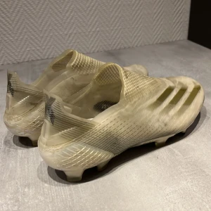 Vita fotbollsskor från Adidas X. Elite sko/ Adidas högsta model - Säljer ett par vita fotbollsskor från Adidas med en stilren design och diskreta ränder på sidan. Skorna har en strömlinjeformad form utan snörning för en tight passform och optimal bollkontroll. Perfekta för spel på gräsplaner.