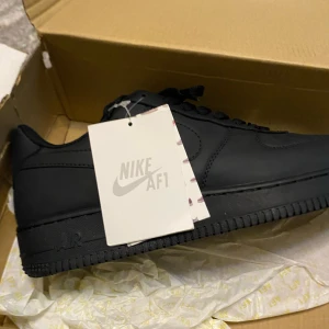 Svarta Nike Air Force 1 - Snygga och klassiska svarta Nike Air Force 1 sneakers i skinn. De har en stilren design med snörning och den ikoniska Nike-loggan på sidan. Perfekta för en casual look. Den är helt ny köpte den och använde 4 gånger och jag vill sälja dom för dom är lite stora för mig 